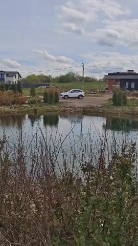 The video pans across a tranquil scene with a pond in the foreground. The water reflects the cloudy sky above. Bushes and trees line the edge of the pond. Beyond the pond, there is a dirt road with a white SUV parked on it. Several modern houses are visible in the background. The camera then moves to show a dirt path running alongside the pond. Rocks and yellow flowers are scattered along the path. The houses in the background remain visible.