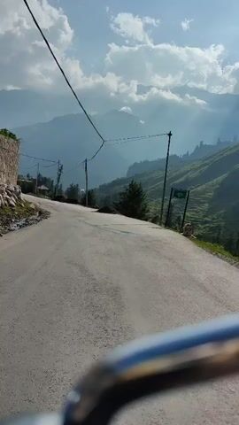 The video shows a view from a vehicle moving along a paved road in a mountainous region. The road curves to the left. On the left side of the road, there is a steep, grassy slope with some trees and a few houses. On the right side, there are more trees and steep, terraced hillsides, likely farmland or pastures. In the background, large mountains rise under a cloudy sky with shafts of sunlight breaking through. Power lines are visible stretching across the scene, supported by utility poles. The foreground is dominated by the vehicle's windshield or handlebars, with a metallic gleam. The overall impression is of a scenic drive through a rural, mountainous landscape.