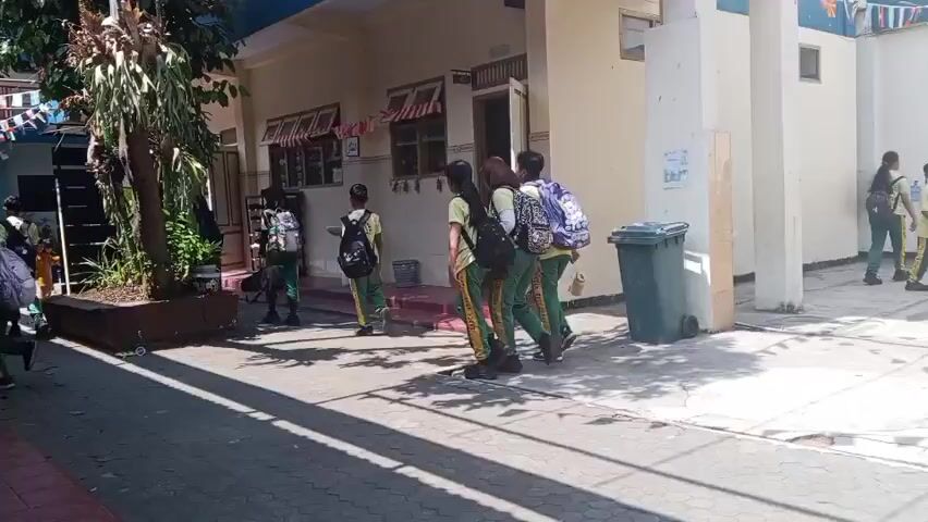 A group of students in matching green and yellow uniforms with backpacks are walking in a courtyard. They are walking towards a building with open doors. There are trees and decorative flags in the courtyard. A trash can is visible on the right side of the image.