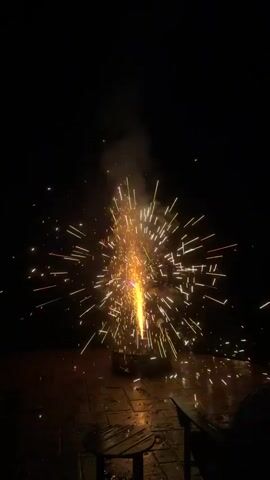 The video shows a fireworks display at night. A firework is lit and erupts with a bright white light and sparks that spray outward. Smoke billows into the air. The fireworks are set off on a paved patio area with two adirondack chairs and a small round table visible in the foreground. The background is dark with some foliage visible on the right.