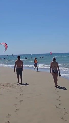 The video shows a sandy beach on a clear, sunny day. The ocean is visible with waves and several people are engaged in water sports, likely kitesurfing or windsurfing, as evidenced by the large kites and sails in the water. The beach is populated with several individuals walking along the shoreline and sitting or standing near the water. In the foreground, a person walks away from the camera on the beach. To the right, another person walks along the shore. In the mid-ground, another person walks towards the water, and further out, a woman in orange shorts walks along the water's edge. More people are seen scattered on the beach and in the ocean, with several colorful kites and sails in the air and on the water. The overall scene depicts a lively beach environment with various recreational activities.