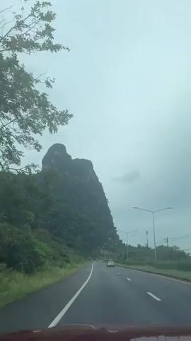 The video captures a view from a moving vehicle on a road. On the left side of the road is a large, forested mountain with a distinctive, somewhat heart-shaped peak. The mountain is covered in lush green trees. The road itself is asphalt, with a white line marking the lane. There are streetlights visible along the right side of the road. A few cars are driving on the road ahead and in the distance. The sky is overcast and appears to be cloudy, suggesting potentially gloomy weather. The overall scene depicts a scenic drive through a landscape dominated by the imposing mountain and the highway.