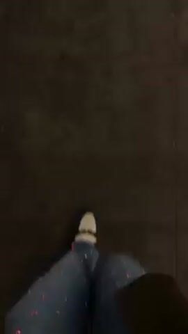 A first-person perspective shot shows a person walking on a dark surface, possibly a sidewalk or pavement. The person is wearing blue jeans with multicolored floral or star-shaped embellishments, and white sneakers. The lighting is dim, obscuring details of the environment. The camera is slightly shaky due to the motion of walking.