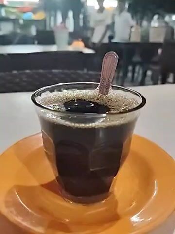 The video shows a clear, geometric glass of dark black coffee sitting on a shiny, bright orange saucer. A small metal spoon rests on top of the coffee. The table surface is white. In the background, out of focus, are people, tables, and chairs, suggesting the setting is a cafe or restaurant, possibly outdoors. The lighting appears soft and even, indicating it might be evening. The camera remains static, focusing sharply on the coffee.