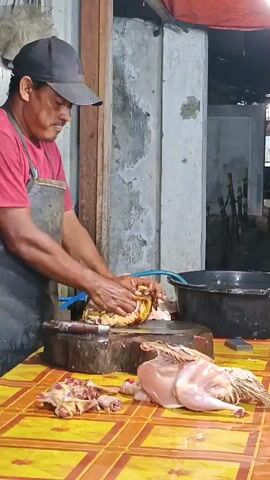 A man wearing a red shirt and a black apron is butchering a chicken at a market stall. He is standing behind a wooden butcher block and using a knife to cut up the chicken. There are several pieces of chicken on the table in front of him, as well as a whole raw chicken. The stall is open-air, and there is a tarp overhead to provide shade. The man appears to be an experienced butcher, and he is working quickly and efficiently.