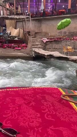 The video shows a fast-flowing river with several outdoor restaurant seating areas set up alongside it. The camera pans from one table with a red tablecloth to show other tables with colorful coverings, and some with cushions on the floor.  The river rushes between and past the seating areas. People are seen walking around in the background, and cars are visible on the hillside. The sky is overcast, and there are trees and buildings on the banks of the river. Sunglasses are visible on the table in the foreground.