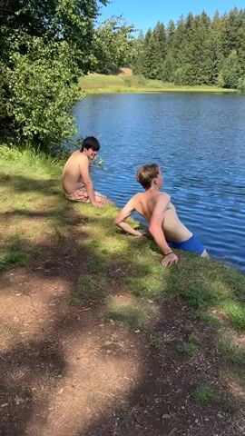 Two young men are sitting on a grassy bank next to a dark body of water. The man on the right is partially submerged in the water, with his legs extended into the lake. He is wearing blue swim trunks. The man on the left is sitting with his back to the camera, wearing checkered swim trunks. The background shows trees and a clear sky. The man on the right turns to look at the camera and states that he has reached the bottom.