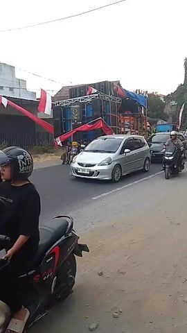 The video shows a street scene with several vehicles and a large sound system setup on the side of the road. In the foreground, a black scooter with two people on it is parked. The person in the back is wearing a helmet with a graphic design, and the person in front is wearing a black t-shirt. The scooter has the word "SPORT" written on its side. To the right of the scooter, a silver Honda Jazz is driving on the road. Behind the Honda Jazz, a black SUV is also on the road. Further down the road, a red motorcycle with a male rider is moving, followed by a black scooter with a male rider. In the background, there is a large structure with many speakers stacked on it, decorated with red and white banners, suggesting a public event or celebration. The environment appears to be outdoors, with a cloudy sky. The overall scene depicts traffic on a road adjacent to a venue with a prominent sound system.