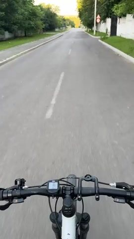 The video is a first-person perspective of someone riding a bicycle on a paved road. The handlebars of the bicycle are visible at the bottom of the frame, with a digital display showing a speed of 16. The road is mostly empty, with trees lining both sides. There are houses and fences on the right side of the road, and a speed limit sign indicating 30 is visible. The sun is shining, suggesting it is daytime, and the sky is clear. The bicycle appears to be moving at a moderate pace.