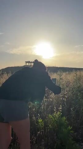 The video opens with a woman in a dark jacket and shorts standing in a field of tall, dry grass. Her back is to the camera, and she is holding a smartphone up to take a photo of the bright sun on the horizon. As the camera shifts, wind turbines are visible in the distance on a ridge. The camera then moves to show a closer view of several wind turbines alongside a dirt road. The sky is blue with scattered clouds.
