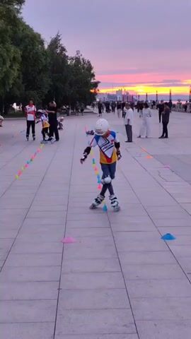 A young person on rollerblades, wearing a helmet, elbow pads, and knee pads, is navigating a slalom course marked by colorful cones. They are in the foreground, facing away from the camera with their right arm extended. In the background, several other people are also on rollerblades, practicing in a line. Some adults are standing and watching. The scene takes place on a paved walkway next to a grassy area with trees. The sky is a vibrant mix of pink, orange, and purple hues, indicating either sunrise or sunset, with a body of water and distant buildings visible on the horizon.