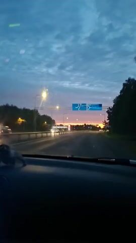 The video shows a view from inside a car driving on a highway at dusk or dawn. The sky is a mix of blue and pink clouds. There are streetlights and overhead signs. The road appears to be a multi-lane highway with trees and buildings visible on the sides. The dashboard of the car is visible in the foreground, and there is a small figurine on the dashboard. The car is moving forward, indicated by the shifting perspective of the scenery.