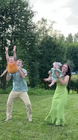 A family is playing in a grassy yard with trees in the background. A man on the left is holding a young boy in front of him. A woman on the right is holding a baby in front of her. The man and woman then lift the children into the air. The children are smiling and laughing. The family is having fun together.