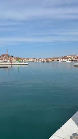 The video pans from a view of a harbor to a marina with boats. The harbor, lined with colorful buildings and crowned by a distant church steeple, reflects off the calm water. As the camera moves, it reveals several sailboats docked at a marina, their masts standing tall against the backdrop of a treeline. The water's surface is smooth, mirroring the boats and the sky. The day appears sunny with a few wispy clouds visible overhead.