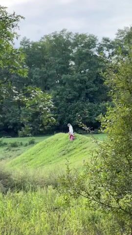 A person in a white top and pink skirt is visible from behind, walking away from the camera on a grassy hill. The person is partially obscured by green leaves from a tree in the foreground. Another person, mostly obscured by the hill, is seen sitting on the hill. The environment is outdoors with lush green grass and trees.