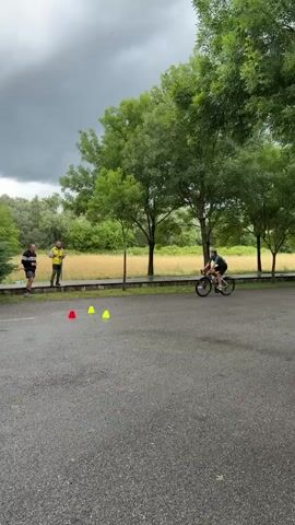 The video shows a cyclist maneuvering around traffic cones in an asphalt parking lot. The cyclist, wearing a black helmet, black and dark green cycling attire, navigates between two yellow and one red cone. Two people, one in a yellow vest and the other in a black jacket, stand to the left, observing the cyclist. The scene takes place outdoors, with green trees and bushes surrounding the parking lot. The sky is overcast.