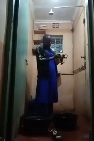 The video shows a person inside a small, simply furnished bathroom. The person is wearing a blue dress and a black jacket with white stripes on the sleeves. They are holding a small object in their hands, examining it closely. The bathroom has a window with bars on it, a small shelf, a sink, and a shower head. Two black plastic tubs are on the floor. The walls are a light peachy color, and the doorframe is painted a greenish-blue. The lighting is dim.