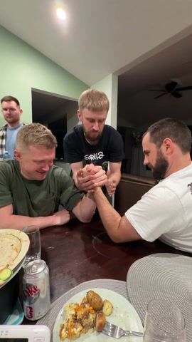 Three men are at a table arm wrestling. One man in a black t-shirt is refereeing. The man on the left in a green t-shirt is exerting himself. The man on the right in a white t-shirt looks more relaxed. There are plates of food, glasses, and a can of Coke on the table. A fourth man can be seen in the background. The room is well lit, with light green walls.