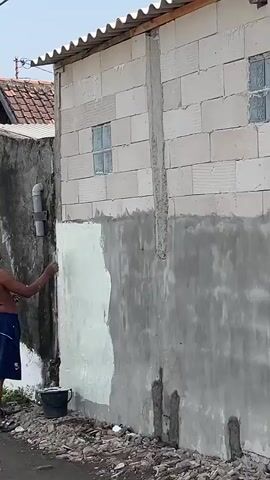 A shirtless man wearing blue shorts applies white plaster to a partially plastered wall with a trowel. He stands in front of a building with a pipe attached to the exterior wall. A bucket sits on the ground near him. The wall has a window made of glass blocks. The ground is covered in dirt and rubble.