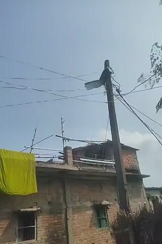 The video displays a series of static images, each lasting approximately one second, depicting an outdoor scene under a clear blue sky. The primary elements captured are buildings, utility poles with wires, and laundry.

In the first frame, a bright yellow garment is prominently displayed, hanging from what appears to be a clothesline or railing on the roof of a brick building. A utility pole with a light fixture and multiple wires is visible to the right. The building is constructed of brick and has at least two windows.

The subsequent frames show a slightly different perspective of the same scene. The yellow garment remains visible in the earlier frames. A tall telecommunications tower can be observed in the distance in some of the later frames. The brick buildings appear somewhat aged, with some visible signs of wear and tear. The overhead wires are numerous and crisscross the sky. The lighting suggests it is daytime, likely with ample sunlight. No people are visible in any of the frames. The overall environment suggests a residential or semi-urban area.
