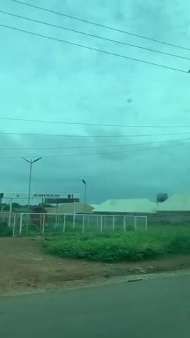 This video captures a moving perspective of a roadside scene in Laminga, Nigeria. The primary subjects are residential buildings with distinctive corrugated metal roofs, varying in completion and style, interspersed with lush green vegetation. Several structures appear to be under construction, indicated by exposed bricks and incomplete walls.

Objects visible include utility poles with power lines crisscrossing the sky, a water tank on one of the rooftops, a rudimentary roadside shelter with stacked tires, a motorcycle, and a white car parked near a building. The scene suggests a community in development, with a mix of established and ongoing construction projects.

The activity depicted is essentially a visual journey along a road, showcasing the landscape and the built environment. There are no clearly discernible people actively engaged in specific tasks, though a figure in yellow appears near a building site, possibly a construction worker.

Notable details include the overcast, greenish sky, which hints at a possibly humid or cloudy day, typical of a tropical climate. The presence of power lines and the style of architecture provide location cues pointing to Nigeria. The overall mood is observational and documentary, offering a glimpse into the everyday scenery of the area. No visible text is present in the video.