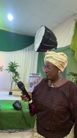 A woman in a brown dress and gold headwrap holds a microphone and sings in front of a stage. The stage has green and white decorations, plants, a light, and a poster. The lighting changes from white to blue and pink. The woman wears glasses and jewelry. She is looking up as she sings.