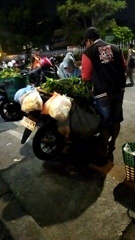 A person in a black jacket and jeans stands by a motorcycle heavily laden with bags and produce. Another person in a hooded jacket is partially visible near the front of the motorcycle. The scene appears to be outdoors at night, with some lighting visible in the background. The motorcycle has a license plate and is parked next to a paved area, possibly a street or parking lot. There are other motorcycles and people in the background.
