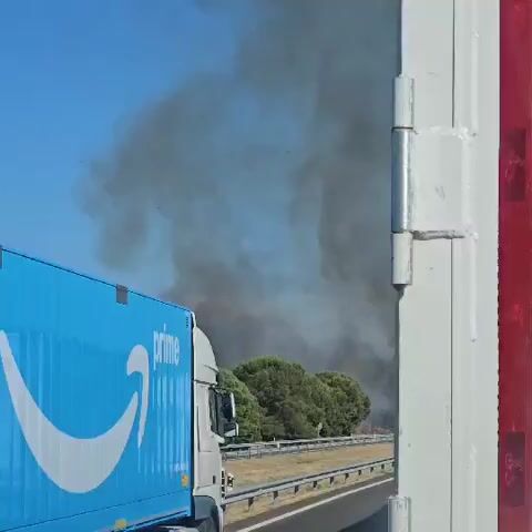 A blue Amazon Prime truck is seen driving on a highway. In the background, there is a large plume of black smoke rising into the clear blue sky, obscuring the horizon behind a line of green trees. The smoke suggests a fire is occurring in the distance.