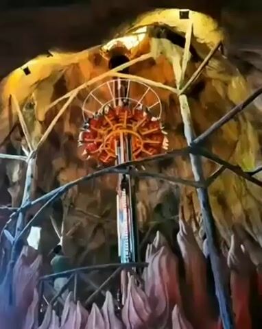 A camera angle looks up at a tall amusement park ride with a circular platform at the top holding people. The ride appears to be in a cave-like structure with rocky walls and scaffolding. The ride is illuminated by warm lighting. The camera zooms out slowly as the ride continues to move.