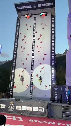 Two competitive climbers ascend a tall, artificial climbing wall divided into two identical vertical sections, each marked with a large circle illuminated by spotlights and lined with numerous red climbing holds. Both climbers are wearing harnesses and athletic clothing. The climber on the left wears a patterned shirt and dark shorts, while the climber on the right wears a bright yellow tank top and dark shorts. Digital timers at the top of each section display the climbers' progress. The competition appears to be taking place outdoors, with a clear sky and mountains visible in the background. Banners and logos, including "WORLD CLIMBING SERIES" and "HBO MAX," are displayed at the base of the wall, indicating a professional sporting event. Spectators are visible in the lower left and right corners of the frame. The video captures the climbers' ascent from near the bottom of the wall to the upper sections.