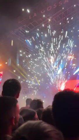 A concert is taking place, likely a rock or pop event based on the stage lighting and effects. Fireworks are being set off in front of the stage, emitting bright white and silver sparks. The crowd is dense, primarily composed of people, visible mainly from the back of their heads, facing the stage. The stage has a large screen with red lighting and a blue image, possibly a logo, displayed. There are spotlights and other lighting elements arranged across the stage structure. The atmosphere is hazy, possibly due to smoke from the fireworks or fog effects. The sign "LA GOSSA" is faintly visible above the stage.