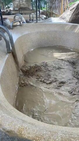 A large, round, speckled tub, possibly made of concrete or fiberglass, is being filled with a slurry of mud or muddy water. A metal pipe is positioned above the tub, and a steady stream of dark, muddy liquid is pouring from the pipe into the tub. The tub is partially filled with this substance, which is churning and swirling as the fresh mud enters. The surrounding area appears to be an outdoor setting, possibly a spa or a similar recreational area, with structures and vegetation visible in the background.