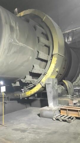 The video shows a close-up, low-angle view of a large, cylindrical industrial machine, possibly a kiln or dryer, within a dimly lit, dusty building. The machine is gray and covered in a fine layer of dust. A yellow metal ring with a segmented design encircles the upper portion of the cylinder. Attached to the side of the machine is a black metal housing with visible bolts and wiring, as well as some yellow pipes or conduits running alongside it. To the right of the main cylinder, there is a wooden pallet stacked with what appears to be metal components, and further to the right, a black metal support structure. The floor is concrete and appears dirty and wet in places. The lighting is low, with some artificial light sources visible, including a white light fixture near the bottom center of the frame. There are no people visible in the video. The overall impression is of an industrial environment that is in operation or has recently been used.