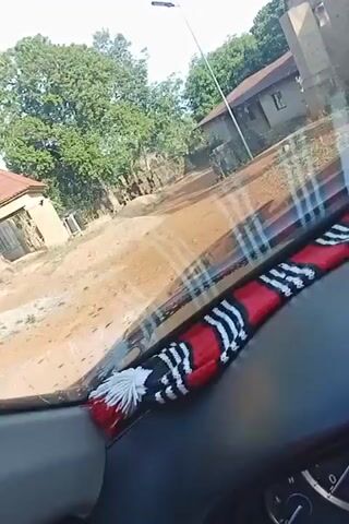 A car’s dashboard with a red, white, and black patterned piece sits on top. The car is driving down a dirt road with buildings on either side. Some of the buildings appear dilapidated. There are trees and a light post. The person in the car states this is their “big home.”