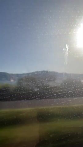 This video is a first-person perspective recording of a scenic landscape viewed through a window. The sun is visible in the upper right corner, casting a bright glare. The foreground is blurred, suggesting motion, and shows green grass and a darker strip, possibly a road or a track. In the mid-ground, there are trees and rolling hills. A body of water, likely a lake or the sea, is visible between the hills. The sky is clear and blue. The video appears to be taken from a moving vehicle, as evidenced by the blur and the changing scenery.