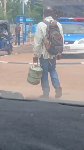A man wearing a light-colored, long-sleeved shirt, jeans, and carrying a dark backpack walks across a paved area, carrying a green metal canister by its handle in his left hand. He is walking away from the camera. In the background, there is a blue and white police vehicle, a three-wheeled vehicle, a person standing near a blue structure, and greenery. The foreground shows the dashboard or interior of the vehicle from which the video is being recorded. The environment appears to be an outdoor, possibly urban or semi-urban area.