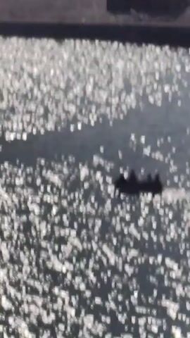 A drone shot flies over a river. The water is dark and choppy, reflecting the sunlight in a shimmering pattern. In the center of the frame, a small, dark boat with several silhouetted figures is visible. The boat appears to be moving across the water. The background shows a dry, rocky landscape with a few scattered structures. The scene is bathed in bright sunlight.