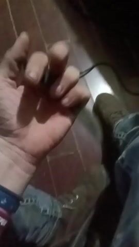 A person's hand is shown from a first-person perspective, manipulating a small object. The hand is positioned in the foreground, with a wooden floor and a person's legs and feet visible in the background. The person is wearing jeans and boots. The object being manipulated appears to be a piece of metal or a tool. The person's fingers move to grip and release the object, and the object is seen to move and spin in their hand. The lighting is dim and appears to be artificial. The overall scene suggests a casual or perhaps preparatory action.