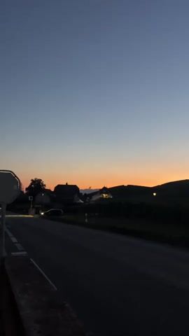 This video shows a rural landscape at dusk. A road curves from left to right in the foreground, with a sidewalk or retaining wall on the left. In the middle ground, there are houses and trees. The sky is a gradient of blue, fading into orange and yellow near the horizon, indicating the sun has recently set. There are vineyards on a hill in the background, with a building that appears to be a church or prominent structure on top of the hill, illuminated by a warm light. The overall scene is peaceful and serene.