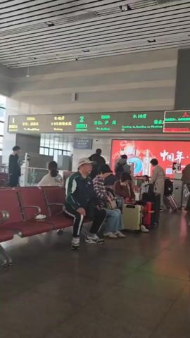 The video shows a waiting area at what appears to be a train station or airport. Several people are sitting on red benches or standing around, many with luggage. Some people are looking at their phones, while others are talking. Large screens display information, including departure times and advertisements. The area has a high ceiling with a striped pattern and large windows providing natural light.  A fan is visible near the window. The scene appears calm and orderly.