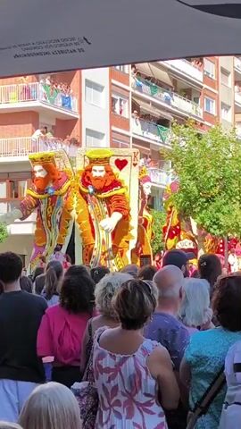 A vertical video captures a street parade with a large float adorned with two figures dressed as king playing cards. The figures are tall, with beards and ornate, colorful costumes in shades of orange, yellow, red, and blue. One figure stands to the left, and the other to the right, both appearing to be holding something in their hands, though it is obscured. Behind them, a large playing card, the king of hearts, is visible. Another smaller float with similarly themed figures and a sign that reads "LUCK" is partially visible to the right. In the foreground, a crowd of people of diverse ages and ethnicities are gathered, facing the float and watching the parade. Some individuals are holding up their phones, presumably to record or take pictures. In the background, brick apartment buildings with balconies are visible, with people watching the parade from the balconies. The day appears sunny with green trees lining the street. The overall atmosphere suggests a festive event.