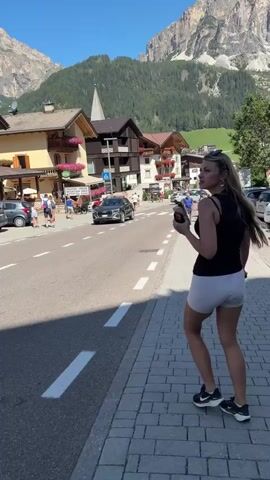 A woman with blonde hair, wearing a black tank top and white shorts, walks on a cobblestone sidewalk next to a road. She is holding a dark object in her right hand and a phone in her left. Cars are parked along the road and some are driving on it. In the background, there are buildings with colorful flowers on their balconies and tall mountains covered in green trees. The sky is clear and blue.