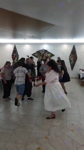 This is a video of a group of people dancing in a room. The room has a tiled floor and is lit by overhead lights. There are decorations on the walls, including some large, dark, spiky structures with floral arrangements. In the foreground, a woman in a pink top and white skirt is dancing, holding a red feather boa. To her left, a man in a plaid shirt and blue shorts is also dancing. Other people are visible in the background, some dancing and some standing. There are tables with what appear to be tablecloths and items on them, possibly food and drinks. The overall atmosphere appears to be a celebration or party.