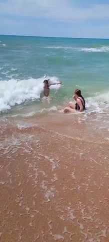 Two young girls in dark-colored swimsuits are playing in the ocean waves near a sandy beach. One girl is sitting in the shallow water, while the other is swimming behind her. The water is turquoise and the waves are white and foamy. The sky is a clear blue. The scene is peaceful and serene.