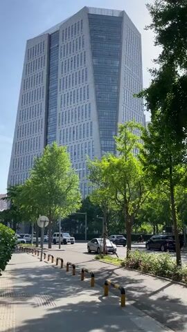 The video shows a street scene with modern buildings, trees, and moving cars. A tall, angular building with many windows stands prominently on the left side of the frame, with the sun glinting off of it. Along the sidewalk are trees lining the street, partially obscuring the building. Cars travel on the road. The sidewalk is lined with short, black and yellow barriers. A flowerbed with small pink flowers sits between the sidewalk and the road. In the distance, a pink apartment building can be seen. People are walking on the sidewalk and through the trees. The sky is clear and bright.