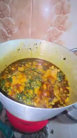 The video shows a pot of soup bubbling on the stove. The pot is silver and has a red base. The soup is a mix of green, orange, and brown colors. There are some leafy greens visible in the soup. The tiles behind the pot are beige with a floral pattern. Steam is rising from the soup, suggesting it is hot and freshly cooked. The camera is held at a slightly high angle, looking down into the pot.