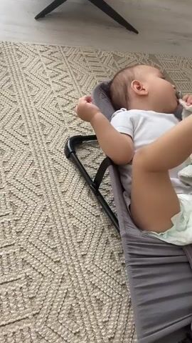 A baby is lying in a gray baby bouncer on a patterned rug. The baby is wearing a white onesie and a diaper. The baby has a pacifier in its mouth and is holding a white and yellow blanket. The baby's legs are raised. In the background, there is a chair, a white curtain, and a toy car.