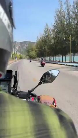 A first-person perspective shot shows a person riding a motorcycle on a paved road with a helmet on. The rider is wearing a green and black plaid shirt. The motorcycle has a digital display showing a speed of 81. There are trees on the right side of the road and water visible behind the trees. A mountain is visible in the distance. Another motorcycle is visible in the distance ahead on the road.
