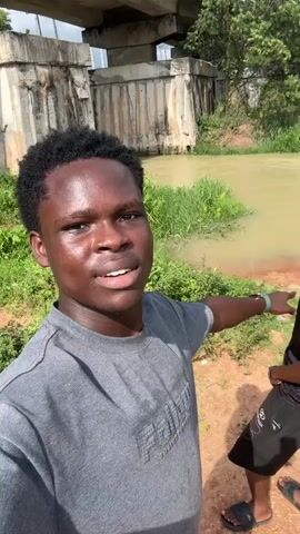 A young man with short, dark, curly hair and a grey t-shirt, possibly with "FEAR OF GOD" embossed on it, faces the camera and speaks, pointing towards a murky, light brown river. He appears to be filming a video, stating, "Come out. This is a river. This river." To his right, partially visible, is another young man wearing a dark t-shirt, dark shorts, and dark slide sandals.

The scene is outdoors, bright and sunny. In the background, a concrete bridge structure spans over the river, which is surrounded by lush green vegetation. The ground is reddish-brown soil. The overall setting suggests a rural or semi-urban area, identified as Ihiagwa, Nigeria, by the provided context. The speaker appears friendly and engaged with the camera.