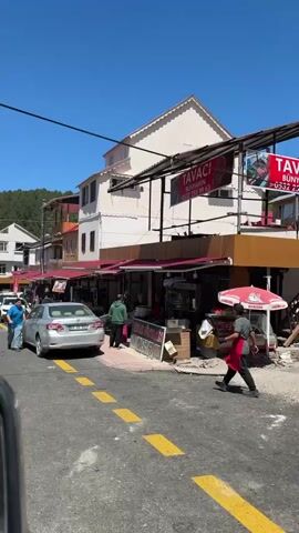 A street scene unfolds with several cars parked and driving on a paved road. On the right side of the road, there are several businesses with awnings and signs. One sign reads "TAVACI KANBEY KEBAB". Another sign displays "KREDİ KARTI GEÇERLİDİR KAVURMA". There are people walking on the sidewalk and in the street. On the left side of the road, there are more buildings and some trees. The sky is clear and blue, indicating a sunny day. The architecture appears to be a mix of traditional and modern styles. The overall impression is a bustling street in a town or small city.